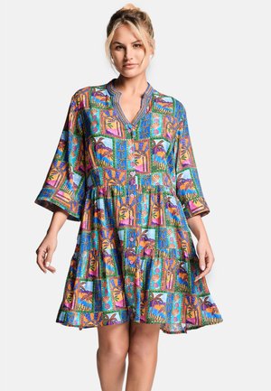 Femme aux cheveux blonds portant une robe colorée, à motifs, longueur genou, manches 3/4 et col en V boutonné, debout devant un fond blanc.