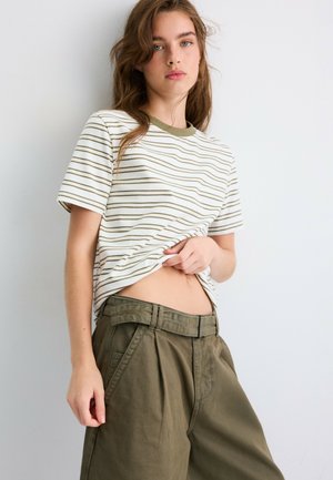 Jeune femme portant un t-shirt blanc à rayures olive relevé pour montrer le ventre, avec un pantalon ample taille haute couleur olive et une ceinture.