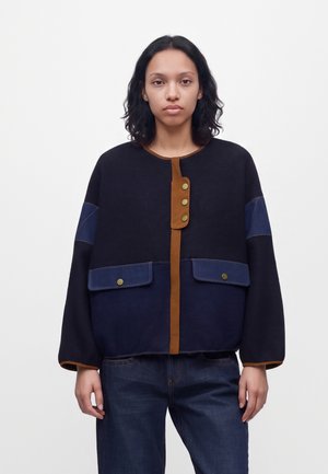 Femme aux cheveux foncés mi-longs porte une veste ample noire et bleu marine avec des bordures marron et des détails de boutons, associée à un jean bleu foncé.