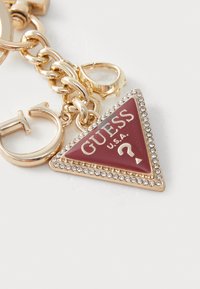 Ciondolo triangolare dorato e rosso con testo "GUESS U.S.A.", bordato da rhinestones trasparenti, attaccato a una catena dorata.