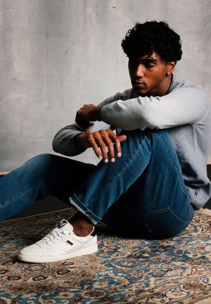 Jeune homme aux cheveux bouclés assis sur un tapis à motifs contre un mur gris, portant un pull gris, un jean bleu et des baskets blanches.