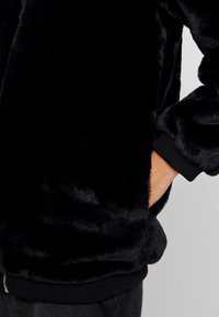 Sort faux fur jakke med blød tekstur, let forlænget krop, ribbede sorte manchetter og en lynlåslomme foran.