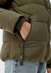 Olive green puffer jacket with quilted design, zippered pocket, and ribbed cuff. Made of smooth, shiny fabric. Casual denim underneath.