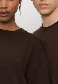 Δύο άτομα που φορούν σκούρα καφέ μπλουζάκια με στρογγυλή λαιμόκοψη, εμφανίζοντας μερικές φορές και τους ώμους τους, το ένα με ένα κρεμαστό σκουλαρίκι αλυσίδα.