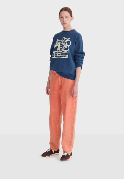 Žena v tmavomodrej mikine s abstraktným béžovým vzorom, voľných vyblednutých oranžových nohaviciach a tmavých teniskách stojaca pred jednofarebným pozadím.