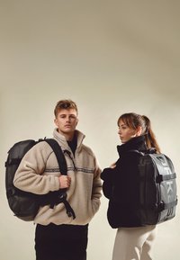 Man och kvinna klädda i fleecejackor och stora svarta ryggsäckar, stående på sidan mot en enfärgad beige bakgrund.