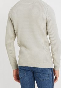 Ljust grå stickad tröja med texturerade ribbmönster, en figurnära design och långa ärmar, tillsammans med blå denimjeans.