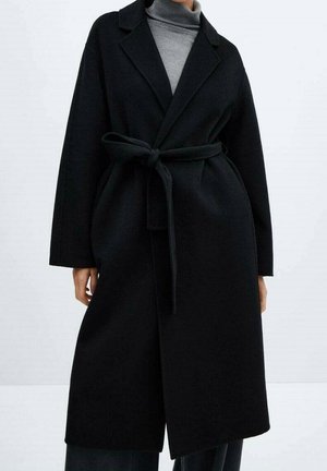 Manteau long noir en laine avec ceinture, porté sur un pull à col roulé gris, associé à un pantalon noir ample, sur un fond clair uni.