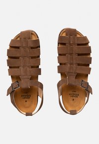 Braune Wildledersandalen mit horizontalen und vertikalen Riemen, offenen Seiten und einer einzelnen verstellbaren Schnalle. Die strukturierte Fußbettung trägt eine Markenkennzeichnung.
