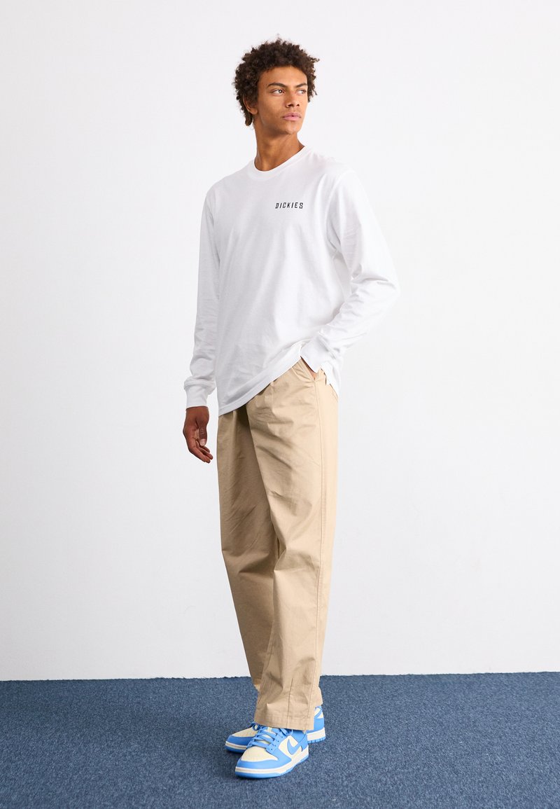 Camisa blanca de manga larga con un pequeño logo negro, pantalones sueltos de color beige y zapatillas azules con acentos blancos. Ajuste relajado y estilo cómodo.
