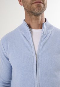 Cárdigan de punto azul claro con cremallera, con un cuello acanalado y textura. Lleva puesto sobre una camisa blanca, con un detalle de cremallera plateada.