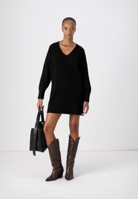 Schwarzes Strickkleid mit V-Ausschnitt, langen Ärmeln und geripptem Material. Kombiniert mit braunen Cowboy-Stiefeln und einer schwarzen Tragetasche.