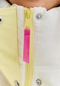 Bunda na zips s hladkým bielym materiálom s vertikálnymi ryhami, žltým zipsom a ružovou stužkou na ťahanie. Sú prítomné strieborné cvoky.