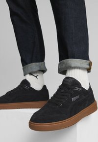 Svarta mockaskor med sula i gummin, med snörningsdesign och Puma-logotyp på sidan. Modellen har på sig uppvikta mörka jeans och vita strumpor.