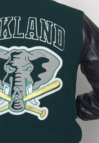 Veste verte avec manches en cuir noir, ornée d'un motif brodé représentant un éléphant et des battes de baseball croisées, avec "OAKLAND" au-dessus.