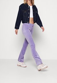 Lavender velour byxkjol med utdragbara ben och dragsko i midjan, kombinerad med en kort vit topp och en mörk denimjacka. Skorna har kraftiga sulor.