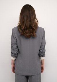 Le blazer gris cintré présente des manches froncées et une texture lisse. Le dos affiche un design simple avec une coupe nette et sans ornements.