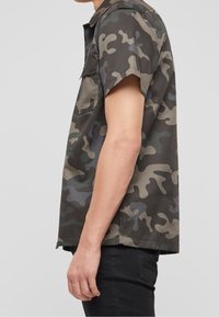 Camicia a maniche corte in camouflage nei toni del verde scuro e grigio, realizzata in tessuto morbido. Presenta una tasca sul petto e una vestibilità rilassata con dettagli minimi.