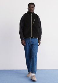 Gilet en polaire noir avec une fermeture éclair à l'avant, associé à un jean en denim bleu clair. Le modèle porte des baskets beiges. Design simple et fonctionnel.