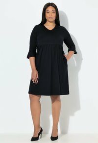 Černé šaty áčkového střihu s tříčtvrtečními rukávy a výstřihem do V. Mají nařasený pas a kapsy na bocích. Vyrobené z měkké látky. Vysoké podpatky.