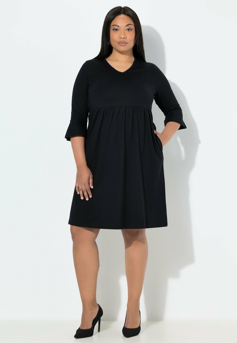 Černé šaty áčkového střihu s tříčtvrtečními rukávy a výstřihem do V. Mají nařasený pas a kapsy na bocích. Vyrobené z měkké látky. Vysoké podpatky.