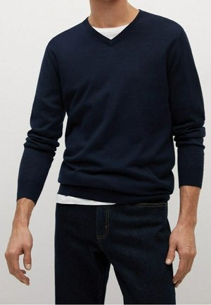Homme portant un pull col V bleu marine superposé à une chemise blanche et associé à un jean bleu foncé, debout avec les bras détendus.