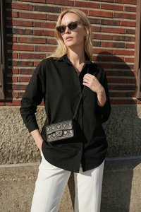 Vrouw draagt een zwart overhemd met knopen, witte broek, zonnebril en een kleine zwarte doorgestikte crossbodytas, staand tegen een stenen muur.