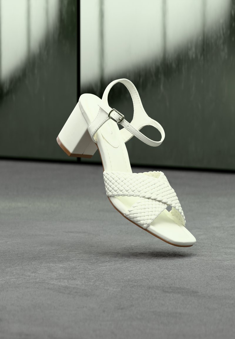 Sandale à talon blanche avec des brides tressées et une boucle à la cheville, posée sur un sol gris devant un arrière-plan réfléchissant flou.