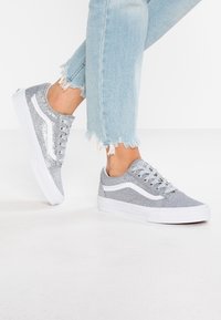 Baskets pailletées grises avec accents blancs, design bas, bout arrondi, six œillets et dessus texturé. Portées avec un jean bleu clair effiloché.