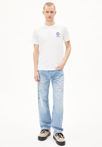 Weißes Baumwoll-T-Shirt mit blauem Sonnenlogo auf der Brust, kombiniert mit hellblauen weiten Jeans und beigen Schuhen mit schwarzen Schnürsenkeln.