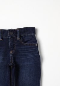 Jeans de denim azul oscuro con bolsillos frontales, trabillas y cierre de botón, mostrados sobre un fondo blanco.