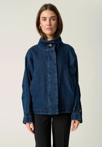 Denim jacket in dark blue with a high collar, gold button closure, and wide sleeves. Features shoulder seams and front pockets.