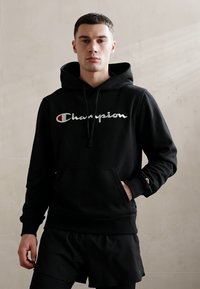 Sweatshirt à capuche noir avec une poche avant, arborant un logo "Champion" blanc et un accent rouge. Tissu doux, coupe décontractée, porté par-dessus des shorts noirs.