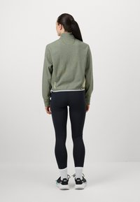 Casaco verde com padrão e fecho éclair, com colarinho alto, usado com leggings pretas. As características incluem uma bainha e detalhes contrastantes.