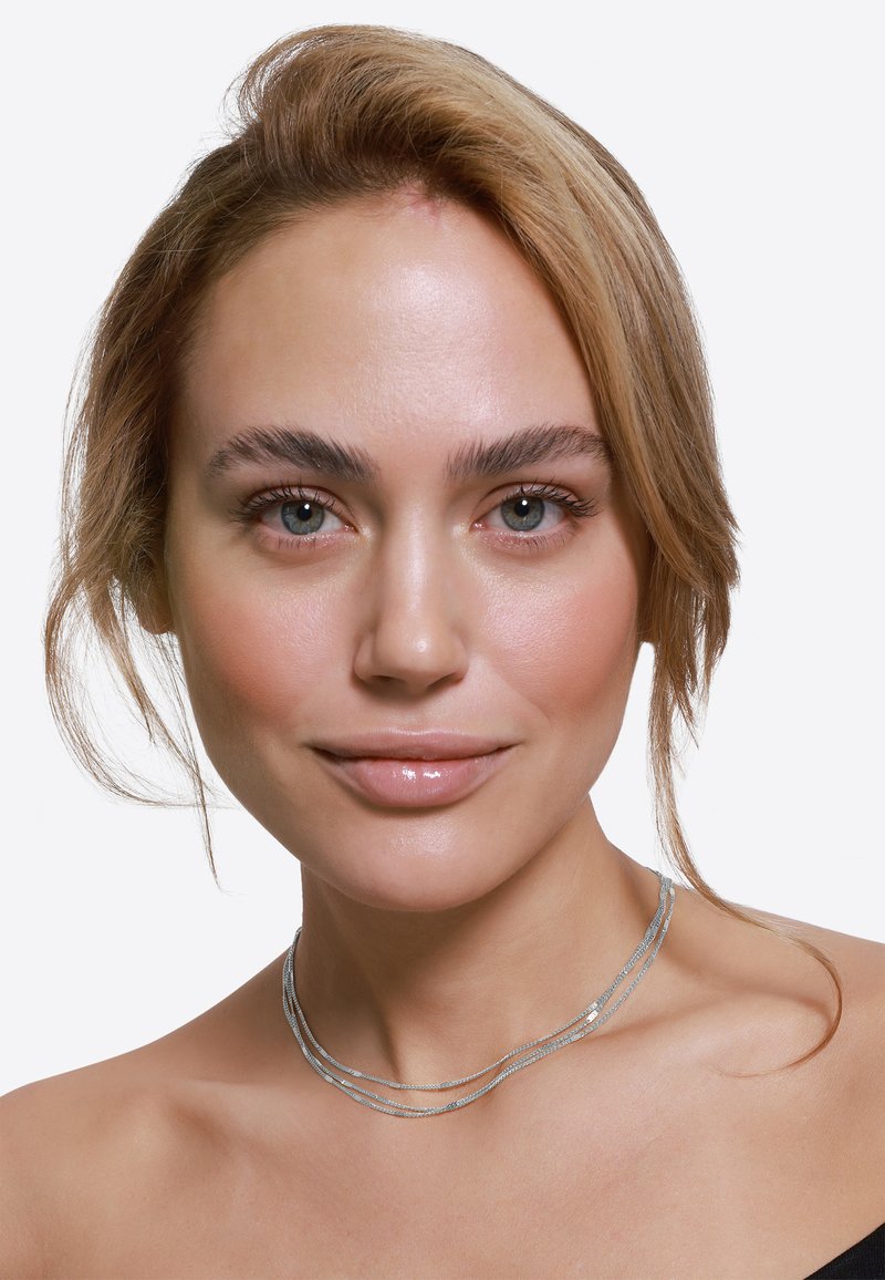 Srebrny naszyjnik warstwowy z cienkimi, strukturalnymi pasmami. Modelka ma jasną skórę i nosi bluzkę z odkrytymi ramionami na neutralnym tle.