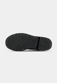 Suela de zapato de goma negra con surcos y crestas texturizadas, que presenta un logotipo rectangular y una marca. Diseño plano y robusto.