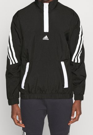 Black Adidas pullover jacket with white zipper detailing and three white stripes on each sleeve, worn by a standing person.