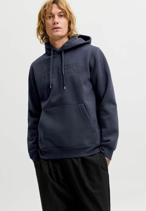 Hombre con el pelo rubio hasta los hombros, vestido con una sudadera azul oscuro Jack & Jones Premium y pantalones negros, de pie frente a un fondo claro y liso.
