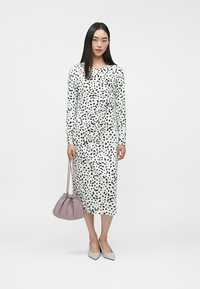 Femme debout dans une robe midi blanche à pois noirs, tenant un sac à main violet, portant des talons blancs sur un fond blanc uni.
