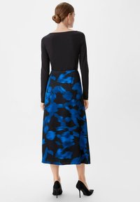 Langärmliges Kleid mit einem schwarzen, figurbetonten Oberteil und einem fließenden Rock mit einem blau-schwarzen abstrakten Muster, ergänzt durch schwarze High Heels.