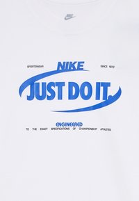 Biele bavlnené tričko s modrým textom: "NIKE," "JUST DO IT," a "NAVRHNUTÉ PODĽA PRESNÝCH ŠPECIFIKÁCIÍ ŠAMPIONSKÝCH ŠPORTOVCOV."