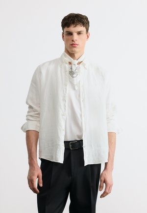Jeune homme portant une chemise blanche à boutons par-dessus un t-shirt blanc, un pantalon noir et un foulard blanc à motifs autour du cou, debout devant un fond uni.
