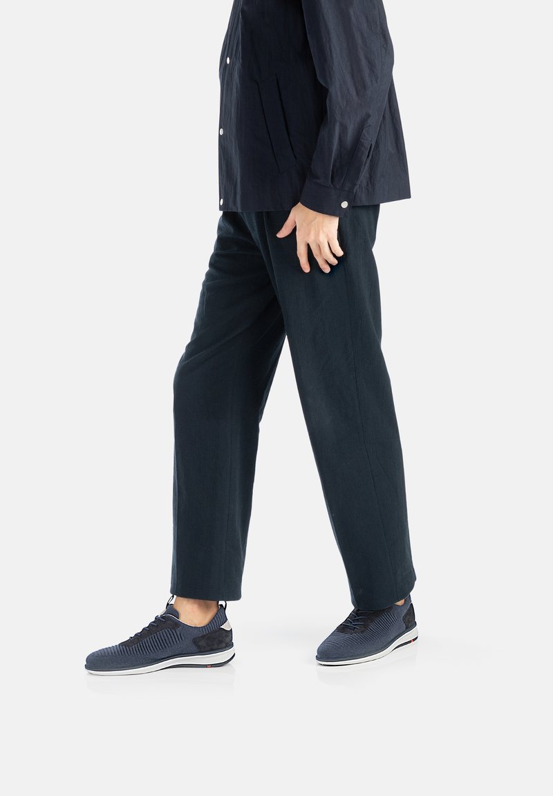 Navyfarbene, strukturierte Sneakers mit Strickoberteil und Wildlederelementen, kombiniert mit lockeren, dunklen Hosen und einem Hemd mit Knöpfen, in Bewegung dargestellt.