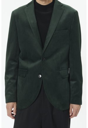 Homme portant un blazer en velours vert foncé avec un seul bouton fermé sur un col roulé noir et un pantalon noir.