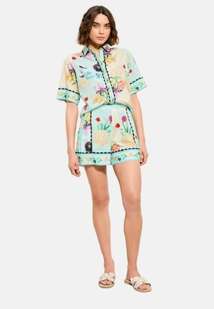 Frau trägt ein kurzärmliges Hemd mit pastellblauem Blumenmuster und passende Shorts, kombiniert mit beigen, geflochtenen Sandalen, steht.