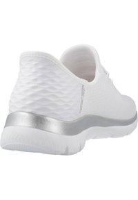 Chaussure de sport blanche avec un dessus texturé et un accent argenté lisse au talon. Elle présente une semelle arrondie et un design de lacet simple.