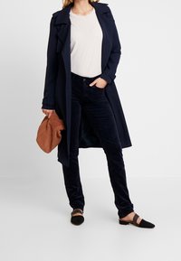 Laivastonsininen trench-coat vaalean yläosan yllä, yhdistettynä tummiin samettihousuihin ja mustiin tasapohjaisiin kenkiin. Ruskea clutch-laukku tuo kontrastia.