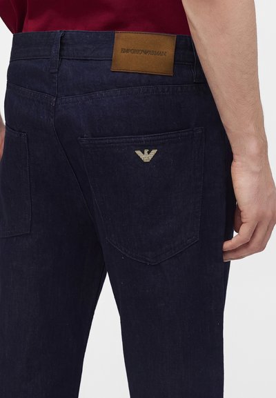 Vaqueros de mezclilla azul oscuro usados por una persona, mostrando bolsillos traseros con logotipos de Emporio Armani y una etiqueta de cuero marrón en la cintura.