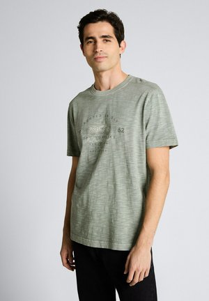 Mężczyzna w jasnozielonej koszulce z krótkim rękawem z nadrukiem i czarnych spodniach, stojący na tle gładkiego jasnoszarego tła.