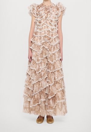 Ärmelloses beige-braunes Maxi-Kleid mit Rüschen und Spitzenbesatz, getragen mit goldfarbenen flachen Schuhen vor einfachem hellem Hintergrund.